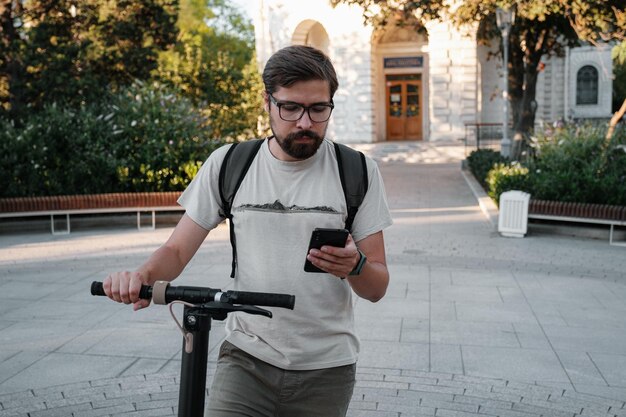 Homem hipster viajante com scooter elétrico ao ar livre na cidade usando smartphone