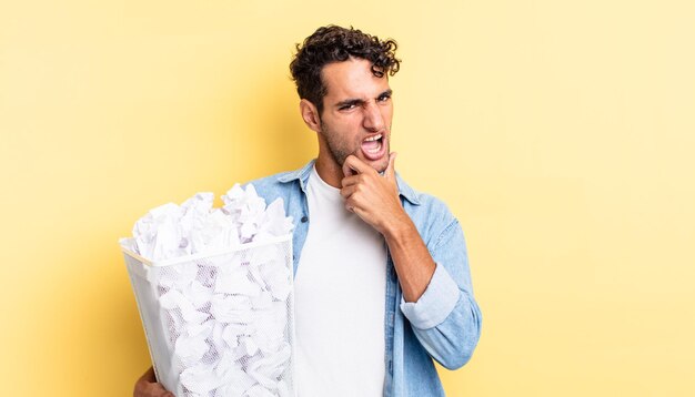 Homem hispânico bonito com a boca e os olhos bem abertos e a mão no queixo. conceito de lixo de bolas de papel