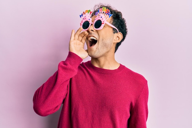 Homem hispânico bonito usando óculos engraçados de feliz aniversário gritando e gritando alto a lado com a mão no conceito de comunicação da boca