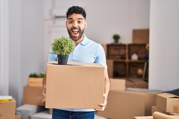 Homem hispânico com barba se mudando para uma nova casa segurando caixa comemorando louco e maravilhado pelo sucesso com os olhos abertos gritando animado.