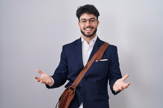 Homem hispânico com barba vestindo roupas de negócios sorrindo alegre de braços abertos como boas-vindas amigáveis saudações positivas e confiantes