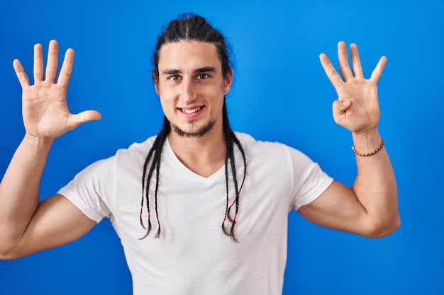 Homem hispânico com cabelo comprido em pé sobre fundo azul mostrando e apontando para cima com os dedos número nove enquanto sorria confiante e feliz