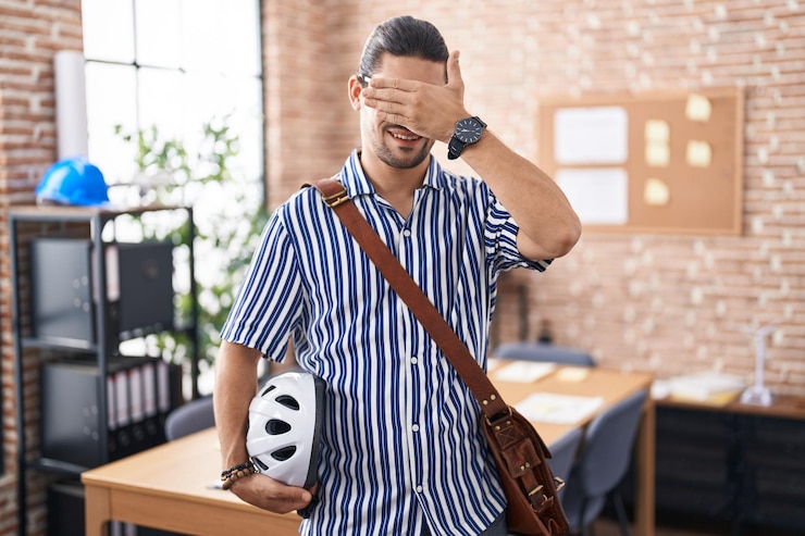 Foto homem hispânico com cabelos longos trabalhando no escritório segurando capacete de bicicleta sorrindo e rindo com a mão no rosto cobrindo os olhos para o conceito cego surpresa