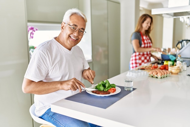 Homem hispânico de meia-idade sorrindo feliz comendo carne com salada enquanto a mulher cozinha na cozinha