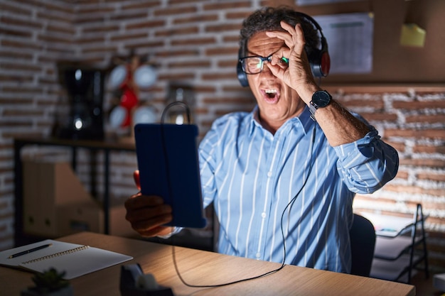 Homem hispânico de meia-idade usando touchpad sentado na mesa à noite fazendo gesto ok chocado com cara de surpresa, olho olhando por entre os dedos. expressão incrédula.