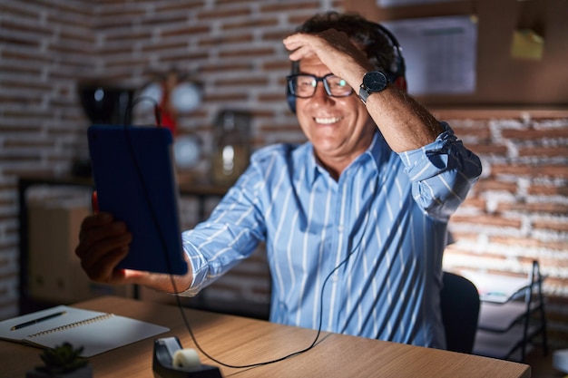 Homem hispânico de meia-idade usando touchpad sentado na mesa à noite muito feliz e sorrindo olhando para longe com a mão sobre o conceito de pesquisa de cabeça