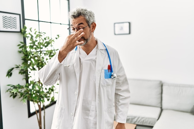 Homem hispânico de meia-idade vestindo uniforme médico e estetoscópio na sala de espera cheirando algo fedorento e repugnante cheiro intolerável segurando a respiração com os dedos no nariz mau cheiro