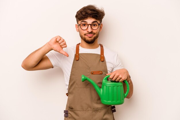 Homem hispânico jovem jardineiro segurando um regador isolado no fundo branco, mostrando um gesto de antipatia polegares para baixo Conceito de desacordo