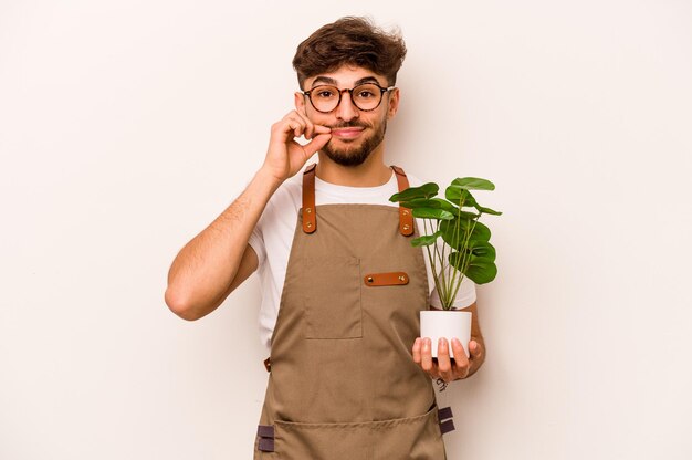 Homem hispânico jovem jardineiro segurando uma planta isolada no fundo branco com os dedos nos lábios, mantendo um segredo