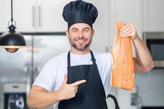 Homem hispânico milenar em uniforme de chef segura salmão de peixe na cozinha cozinhando menu de restaurante de peixe com