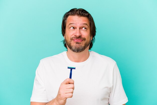 Homem holandês de meia-idade segurando uma lâmina de barbear isolada em um fundo azul confuso, sente-se em dúvida e inseguro.