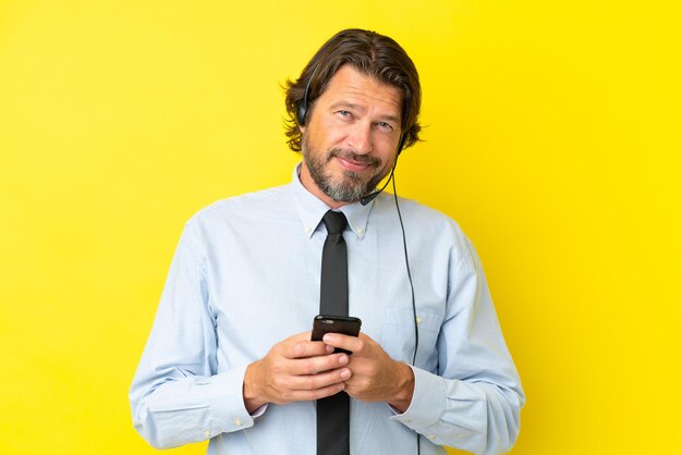 Homem holandês de telemarketing trabalhando com um fone de ouvido isolado em fundo amarelo, enviando uma mensagem com o celular