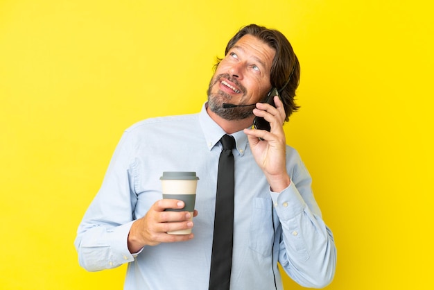 Homem holandês de telemarketing trabalhando com um fone de ouvido isolado em fundo amarelo segurando café para levar e um celular