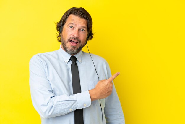 Homem holandês de telemarketing trabalhando com um fone de ouvido isolado em fundo amarelo surpreso e apontando o lado