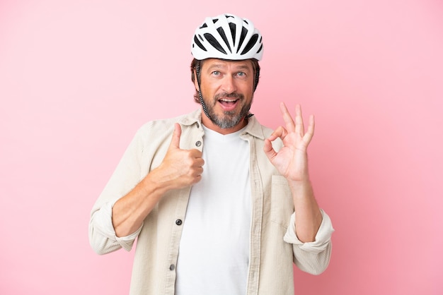 Homem holandês sênior com capacete de bicicleta isolado em fundo rosa, mostrando sinal de ok e gesto de polegar para cima