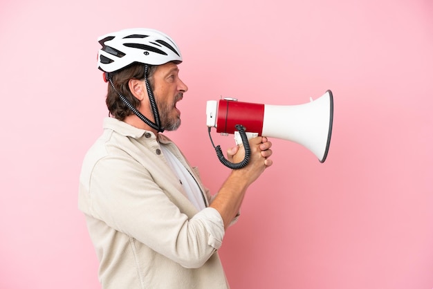 Homem holandês sênior com capacete de bicicleta isolado em um fundo rosa gritando através de um megafone