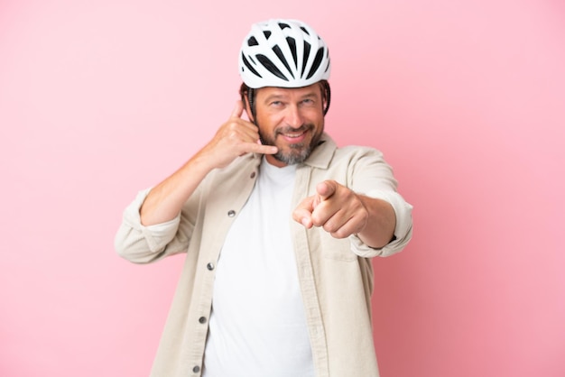 Homem holandês sênior com capacete de bicicleta isolado no fundo rosa, fazendo gesto de telefone e apontando para a frente