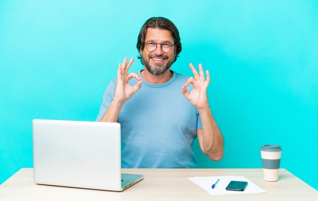 Homem holandês sênior em uma mesa com um laptop isolado em fundo azul, mostrando um sinal de ok com os dedos