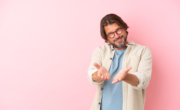 Homem holandês sênior isolado em um fundo rosa segurando copyspace imaginário na palma da mão para inserir um anúncio