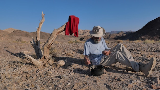 Foto homem idoso a comer e a usar telemóvel no deserto