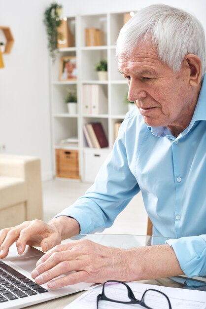 Homem idoso aposentado de camisa azul pressionando as teclas do teclado do laptop enquanto navega na rede para obter dados online em casa