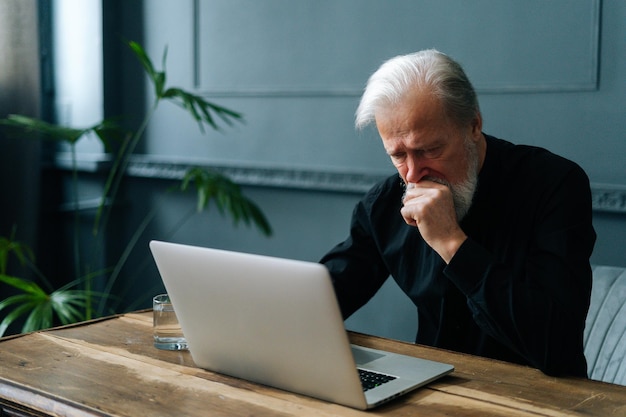 Homem idoso barbudo preocupado deprimido sentado à mesa de madeira com laptop comparar na sala de estar escura