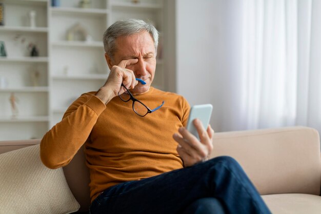 Homem idoso cansado esfregando os olhos usando smartphone em casa