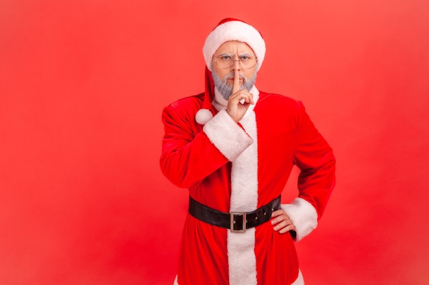 Homem idoso com barba grisalha vestindo fantasia de Papai Noel, segurando o dedo nos lábios, fazendo gesto de silêncio silencioso, pedindo para manter segredo, não fale. Tiro de estúdio interno isolado sobre fundo vermelho.