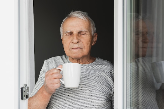 Homem idoso com uma xícara de café quente olhando pela janela aberta