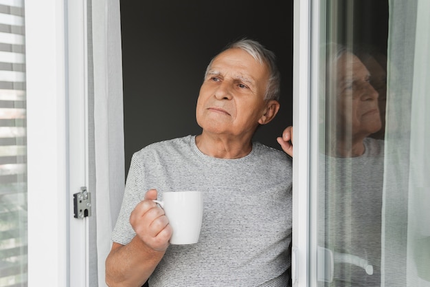 Homem idoso com uma xícara de café quente olhando pela janela aberta