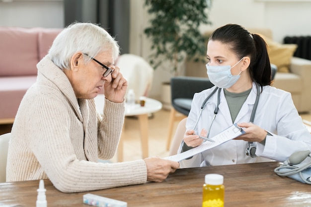 Homem idoso doente olhando um documento médico mantido pela jovem médica sentada à mesa na frente dele durante uma consulta domiciliar