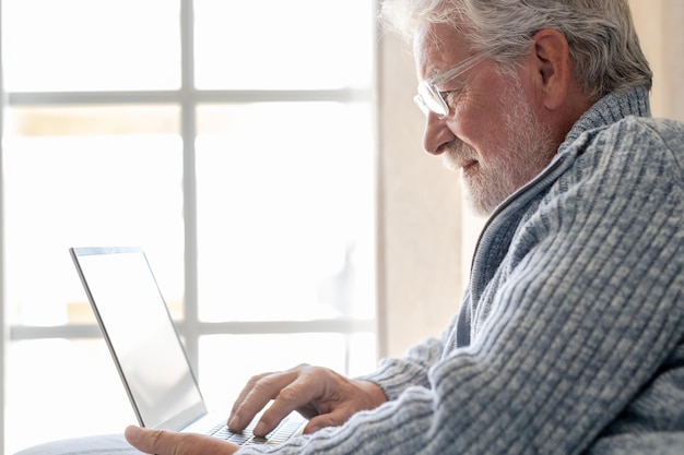 Homem idoso dos anos 70 sentado no sofá navegando no laptop sênior olhando para a tela do laptop navegando na rede geração mais antiga e conceito de uso fácil de aplicativo de tecnologia moderna