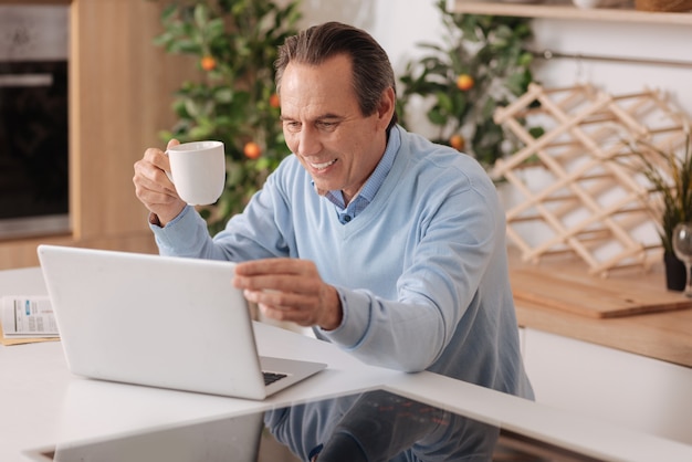 Homem idoso encantado positivo sentado na cozinha enquanto toma uma xícara de café e usa o laptop