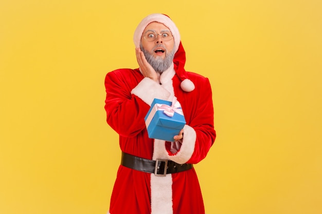 Homem idoso extremamente surpreso com barba grisalha, vestindo fantasia de papai noel, segurando uma caixa de presente azul embrulhada, tem um presente na véspera de ano novo. estúdio interno, tiro isolado em fundo amarelo.