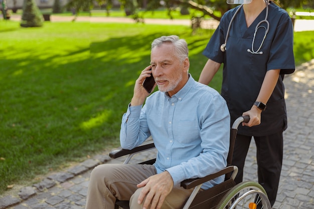 Homem idoso recuperando paciente em cadeira de rodas falando ao telefone enquanto caminhava com a enfermeira no
