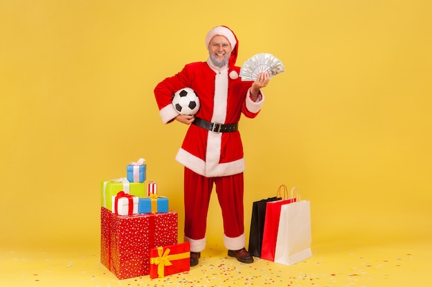 Homem idoso satisfeito feliz vestindo fantasia de papai noel em pé segurando uma bola de futebol e um fã de dinheiro, apostar e ganhar, comprar presentes para o natal. estúdio interno, tiro isolado em fundo amarelo.