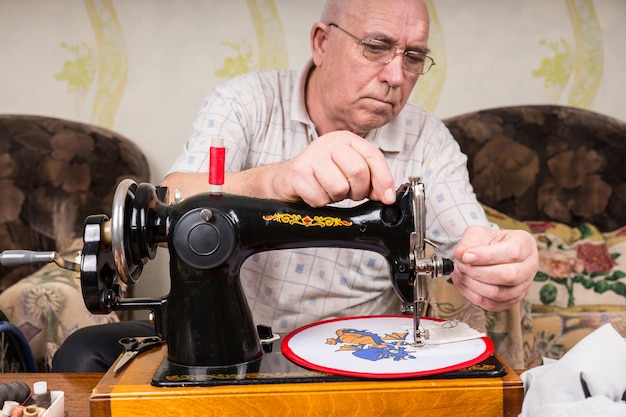 Homem idoso sério usando óculos, trabalhando em uma embarcação de enforcamento de parede com ponta de agulha, usando uma máquina de costura antiquada em casa na sala de estar