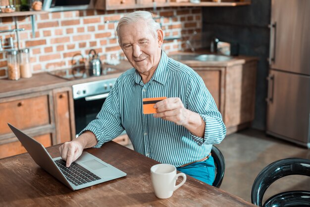 Homem idoso simpático e alegre segurando um cartão de crédito e fazendo um pagamento online enquanto usa seu laptop