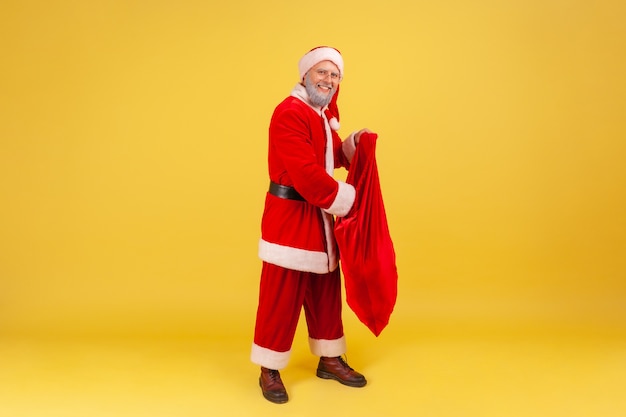 Homem idoso sorridente com barba grisalha vestindo fantasia de Papai Noel, colocando a mão dentro da grande sacola vermelha cheia de presentes de Natal, cumprimentando com o ano novo. Foto de estúdio interna isolada em fundo amarelo