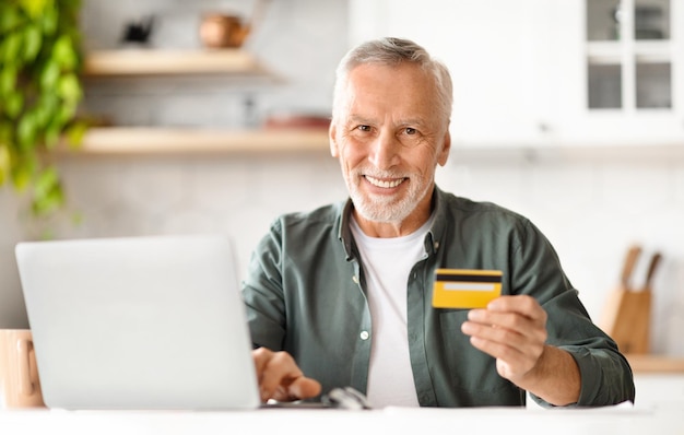 Homem idoso sorridente de comércio eletrônico segurando cartão de crédito e usando laptop em casa