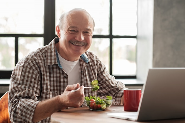 Homem idoso tomando café da manhã na frente de um laptop