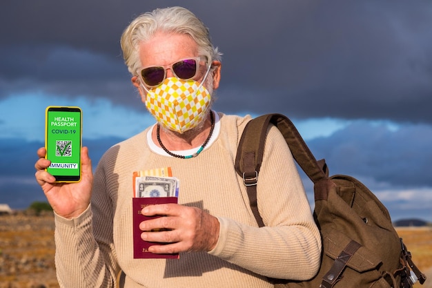 Homem idoso usando máscara protetora no rosto mostra aplicativo de passaporte de saúde para pessoas vacinadas contra coronavírus, prontas para viajar novamente