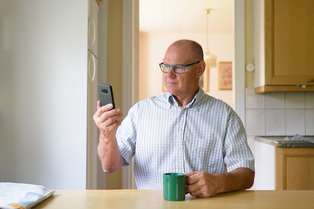 Homem idoso usando o telefone enquanto bebe café perto da janela