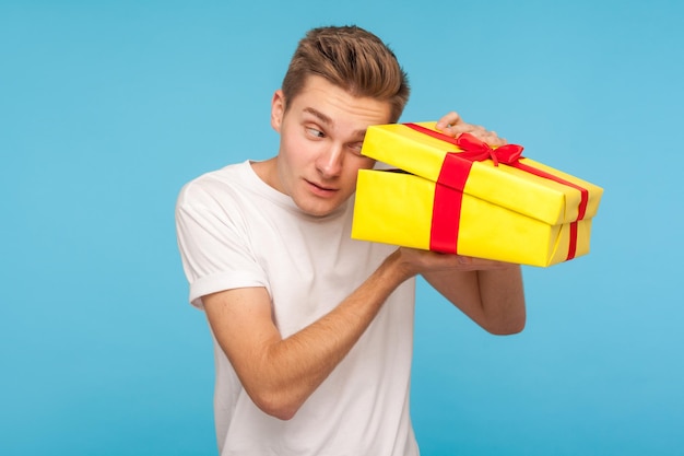 Homem impaciente intrometido em t-shirt branca, olhando para dentro da caixa de presente com curiosa expressão feliz, abrindo o presente antes da celebração, espiando com interesse. tiro de estúdio interno isolado em fundo azul