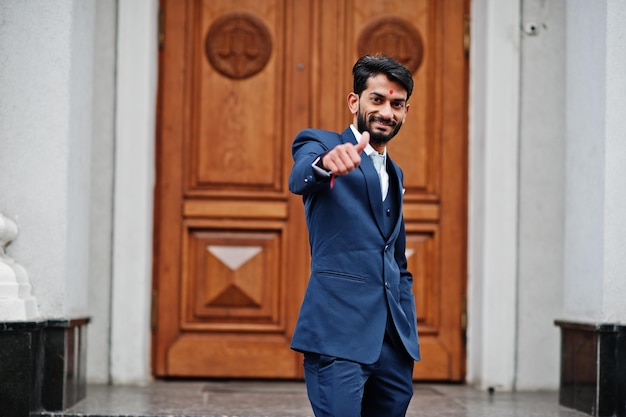 Homem indiano de barba elegante com bindi na testa usa terno azul posou ao ar livre contra a porta do prédio e mostra o polegar para cima