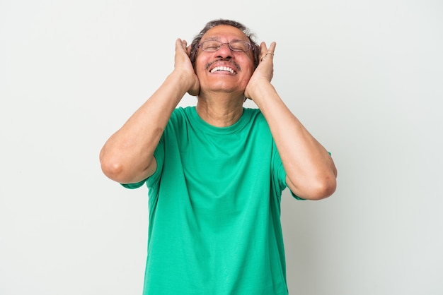 Homem indiano de meia idade, isolado no fundo branco, ri, mantendo as mãos na cabeça com alegria. Conceito de felicidade.