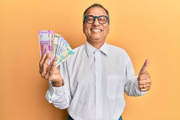 Homem indiano de meia-idade segurando notas de rúpia indiana sorrindo feliz e positivo polegar para cima fazendo excelente e sinal de aprovação