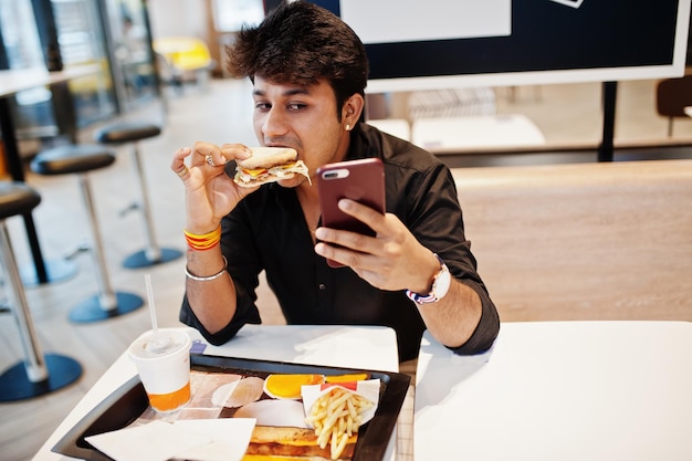 Homem indiano elegante e engraçado sentado no café fast food e comendo hambúrguer e fazendo selfie por telefone