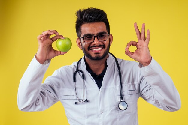 Homem indiano em uniforme médico comendo maçãs no fundo amarelo do estúdio.