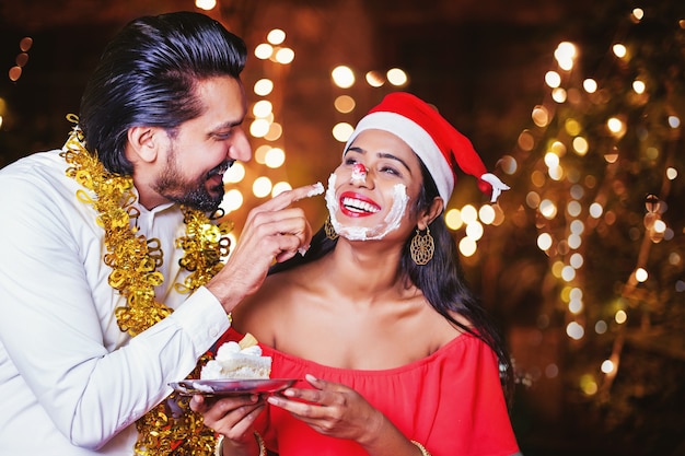 Homem indiano feliz aplicando creme de bolo no rosto da mulher no Natal